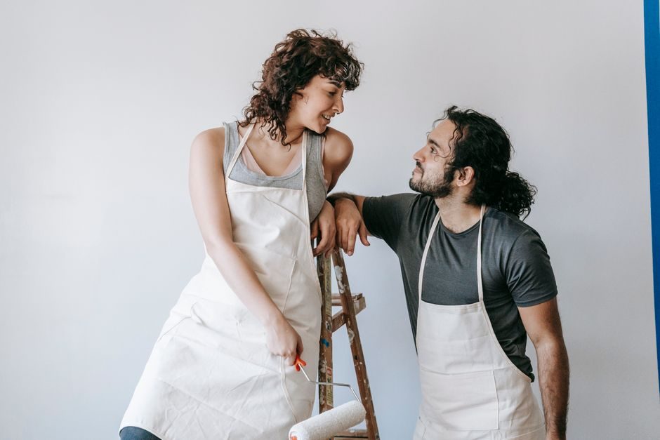 Smiling couple in aprons painting a wall with a roller, enjoying the renovation process.