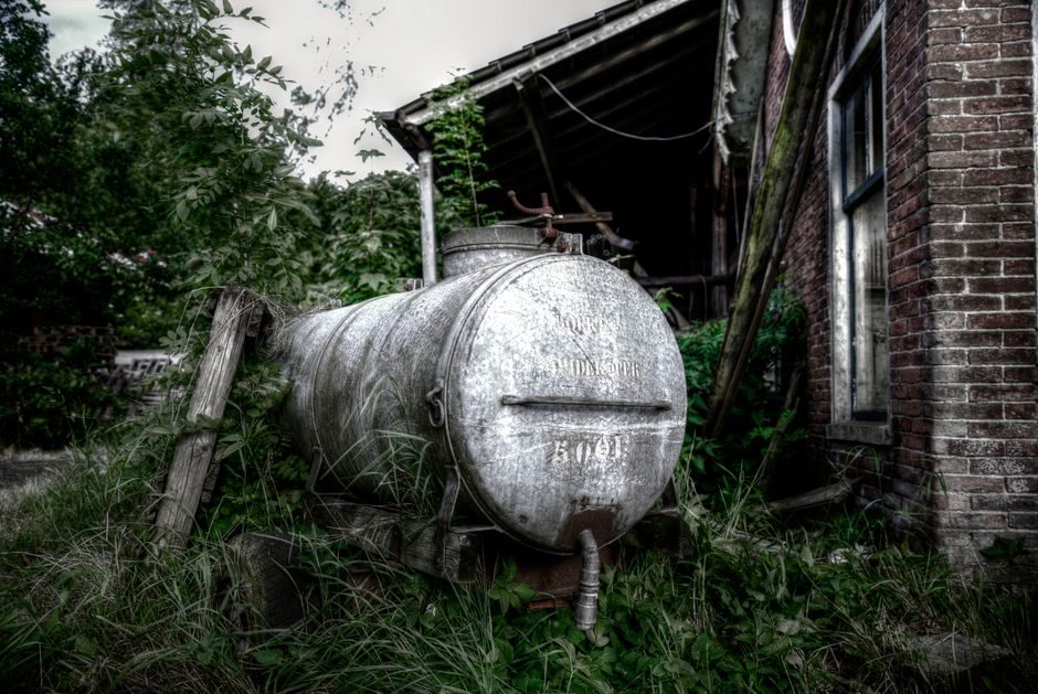 A dilapidated house in a rural setting with a rusted water tank and overgrown vegetation.